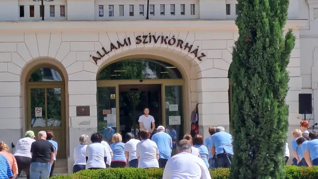 Szívkórház főbejárata, a boltíves bejáratban egy tornát tartó hölgy áll. Előtte egy tuja körül tornázók csoportja