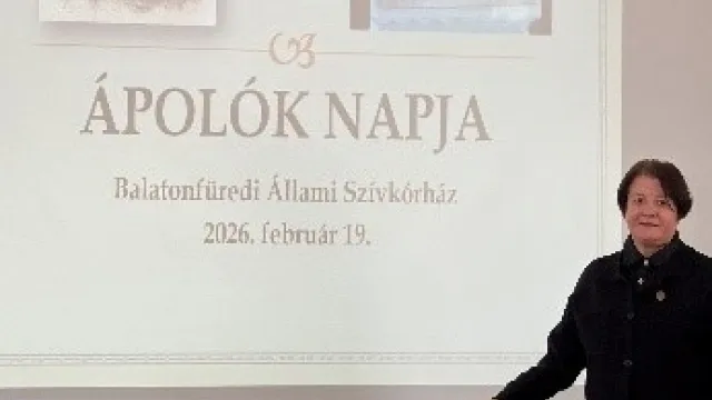 A kép háttere a falra vetített Ápolók napja köszöntő képkockája látszik. A kép alsó részében egy vörösesbarna asztalon egy hatalmas virágcsokor, előtte áll a köszöntőt mondó ápolási igazgató asszony 