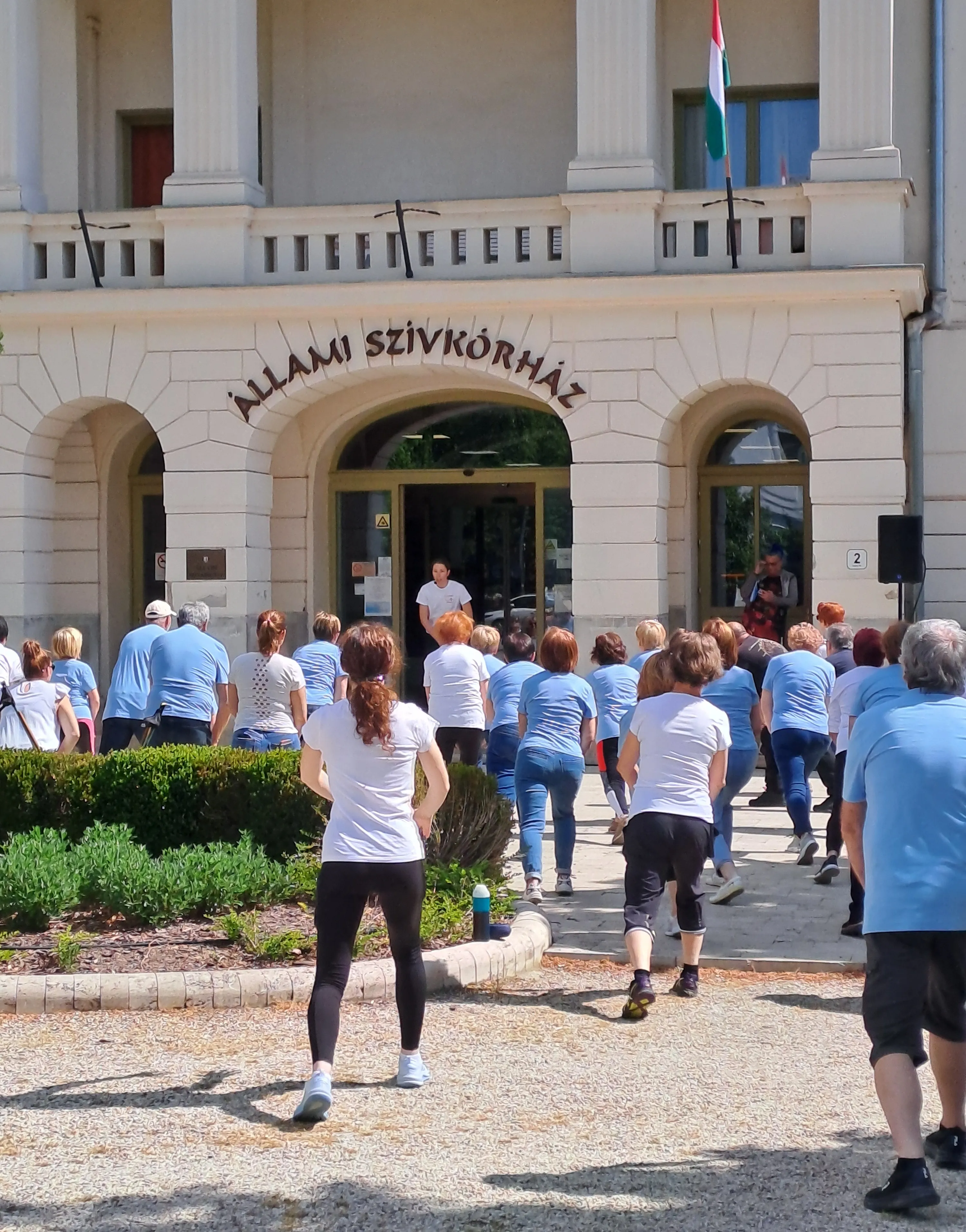 A szívkórház főbejárata, ahol a boltív alatt áll a tornát vezető gyógytornász, előtte a vele tornázó emberek