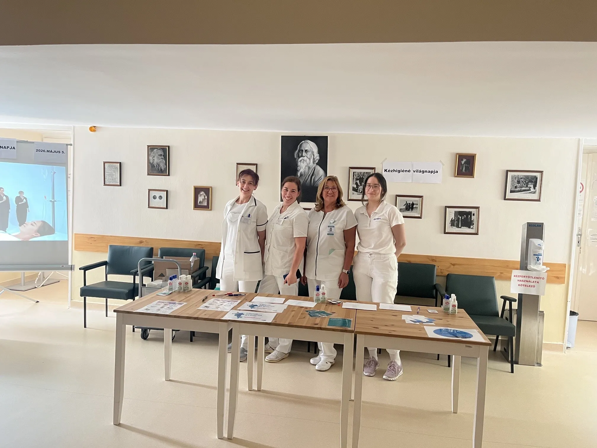 A kép közepén 4 mosolygós kórházi dolgozó hölgy áll. Előttük az asztalon a kézhigiénés vilagnaphoz köthető rejtvények és ajándékok vannak. A hölgyek mögött sötétzöld kórházi székek vannak, a falon pedig Rabindranath Tagore-ról életképek, portrék vannak vannak kitéve.
