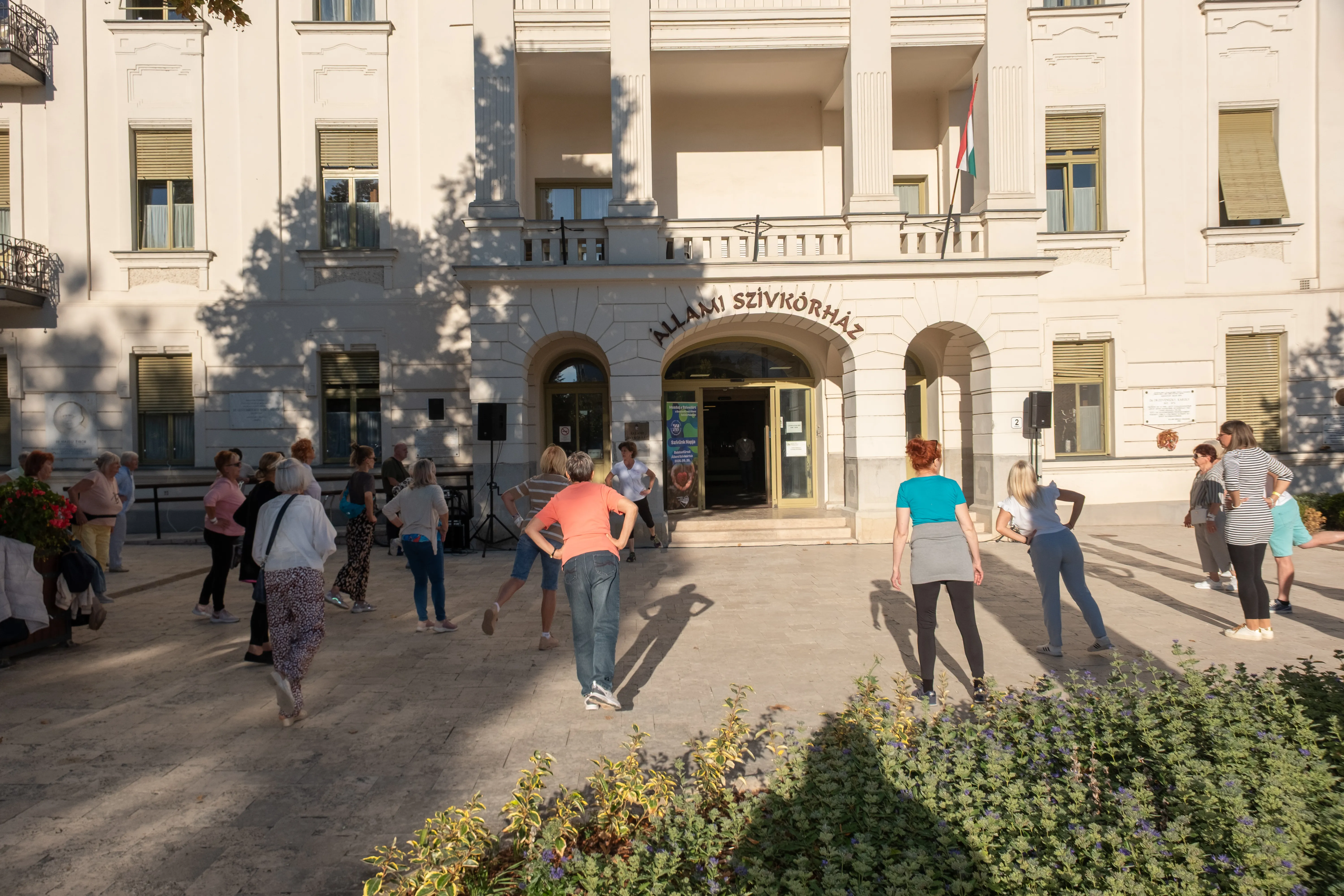 A kórház főbejárata a világos falon Állami Szívkórház felirat. Előtte a tornázók csoportja a tornát tartó gyógytornásszal
